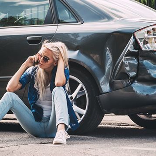 Woman on her phone sitting on the ground next to her damaged car.